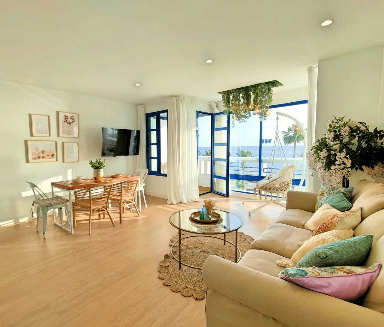 A bright seaside living room with blue window frames, a sofa with sea-glass pillows and the ocean outside.