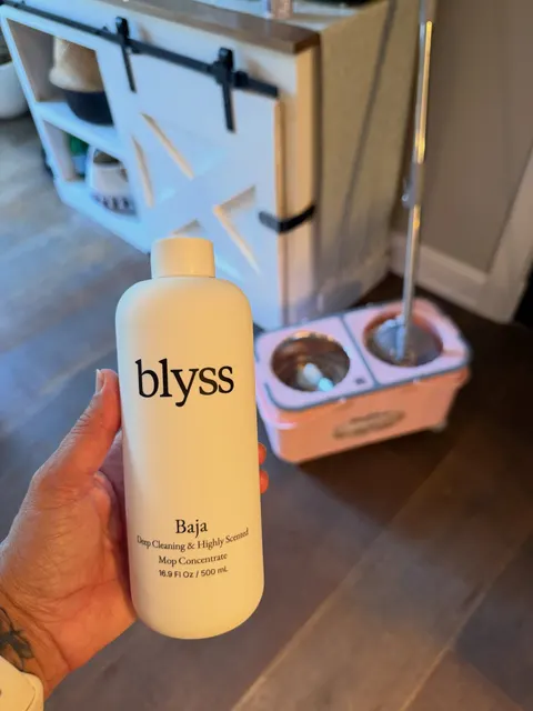 A hand holding a bottle of Blyss Baja mop concentrate beside a spin mop bucket in a bright home.